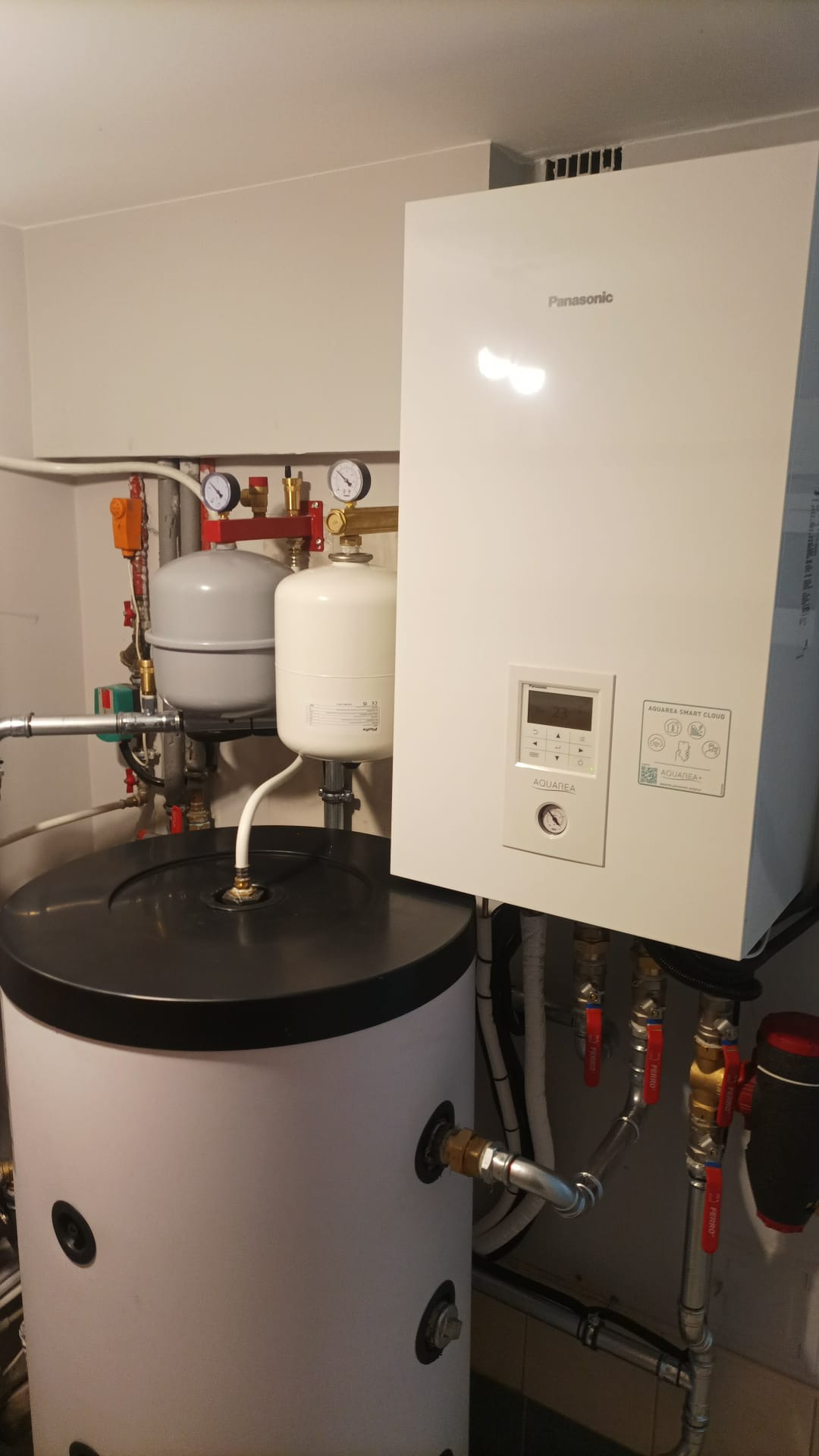 Biała pompa ciepła Panasonic Aquarea Smart Cloud z czarnym zbiornikiem i instalacją hydrauliczną w pomieszczeniu technicznym.