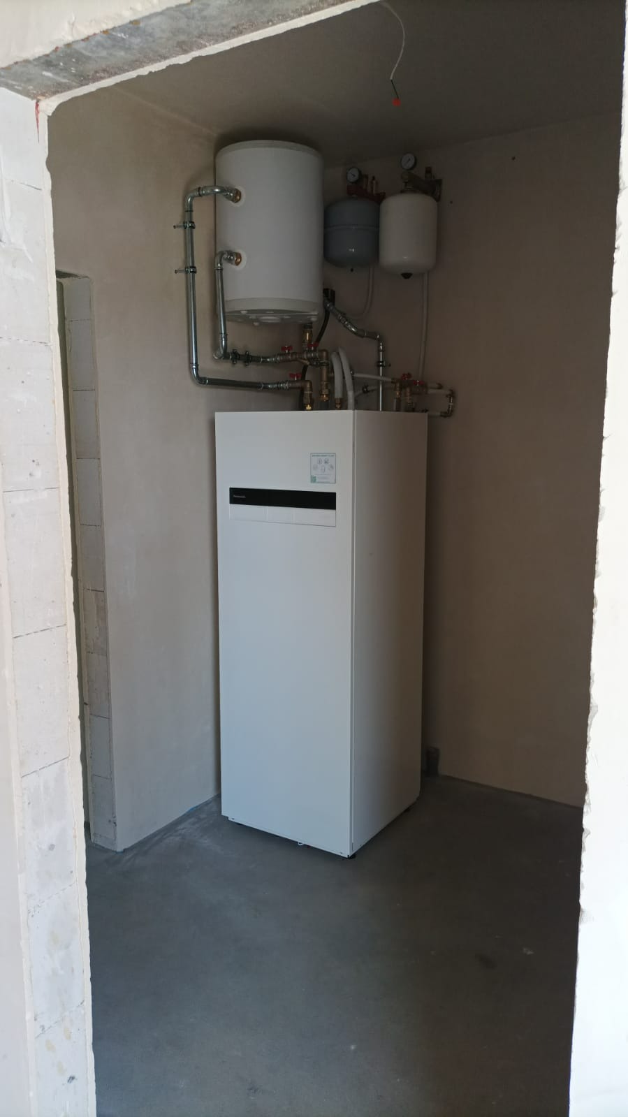 Biała pompa ciepła z zbiornikiem buforowym i osprzętem w surowym pomieszczeniu, widok zza progu. Instalacja grzewcza po montażu, przed wykończeniem ścian.