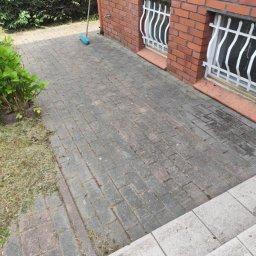 Kamil lisewski - Umyta kostka brukowa przed budynkiem z czerwonej cegły, widoczne okna z białymi kratami i zielony krzew. Na kostce leży niebieska miotła.