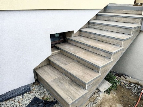 Zewnętrzne schody wyłożone płytkami imitującymi drewno, z szarymi listwami wykończeniowymi. Widoczny fragment elewacji budynku z małym okienkiem piwnicznym.