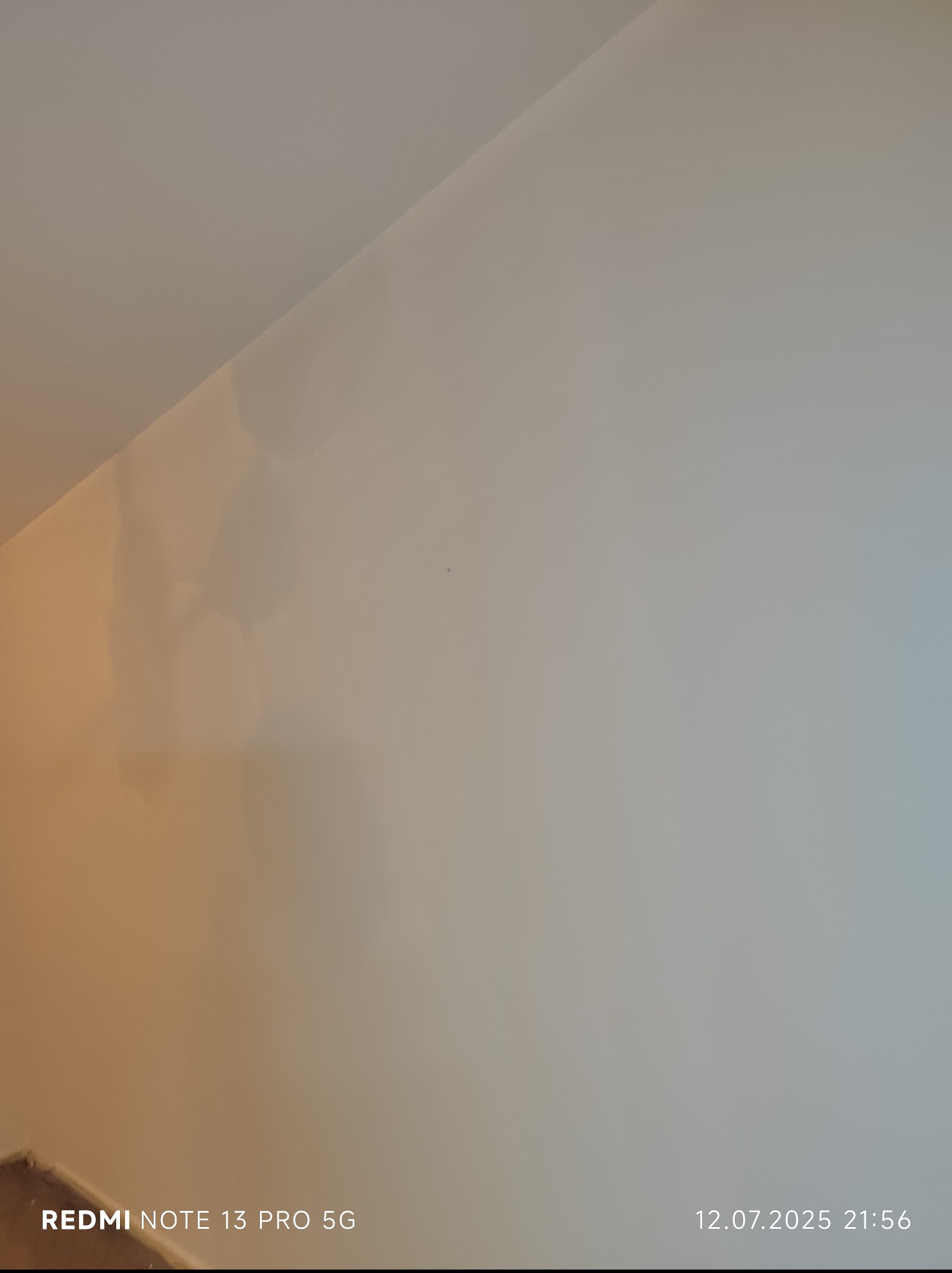 Ściana w odcieniach bieli i beżu pod skosem, oświetlona z lewej strony. Widoczny fragment podłogi i cień. Zdjęcie z telefonu.