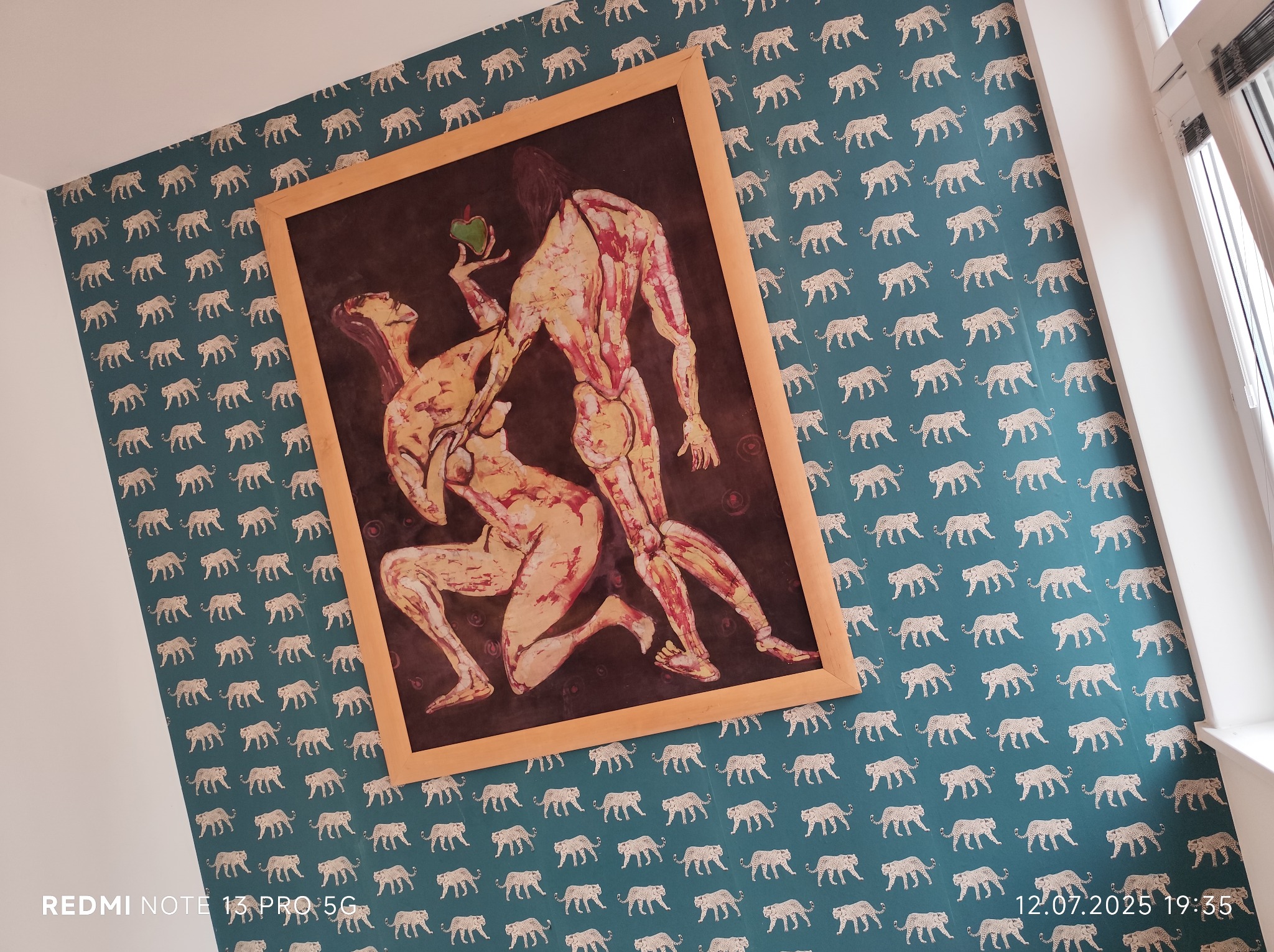 Ściana z tapetą w panterki, na niej wisi obraz w drewnianej ramie przedstawiający akt Adama i Ewy z jabłkiem. Widok na fragment okna z roletami.