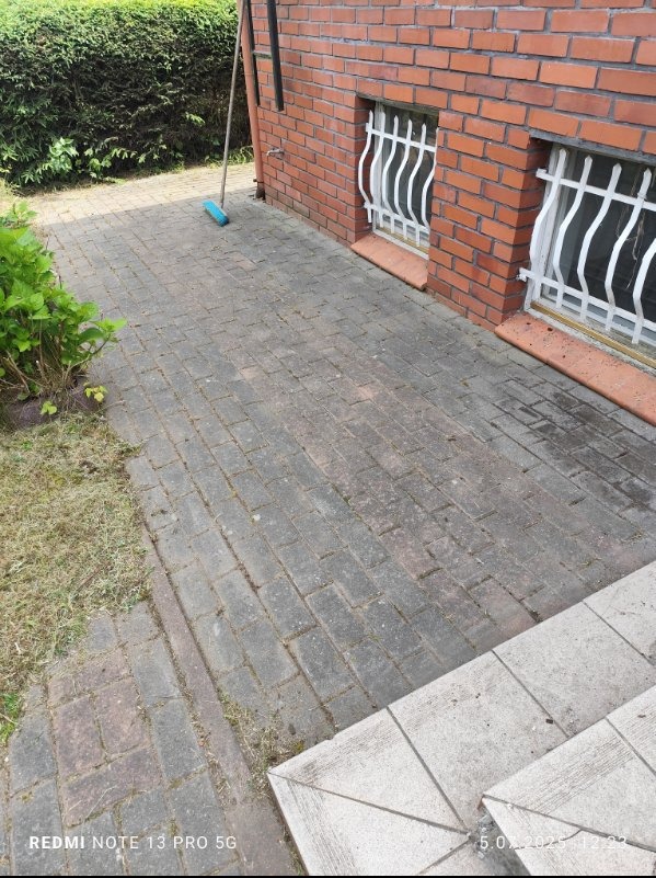 Umyta kostka brukowa przed budynkiem z czerwonej cegły, widoczne okna z białymi kratami i zielony krzew. Na kostce leży niebieska miotła.