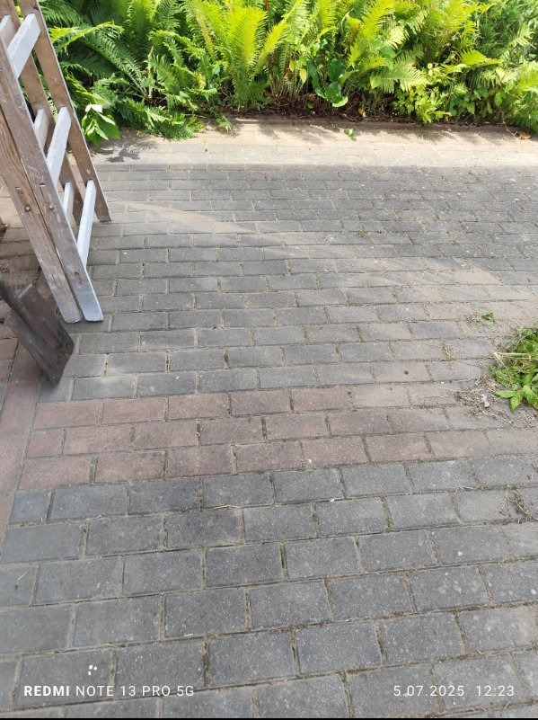 Ułożona kostka brukowa na zewnątrz, z drabinką i zielenią w tle. Widoczny fragment łopaty. Kąt ujęcia z góry, naturalne oświetlenie.