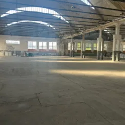 Przestronna hala magazynowa z betonową podłogą i rzędem okien w górnej części ścian. Widoczne słupy nośne i elementy konstrukcji dachu. Dostęp światła dziennego.