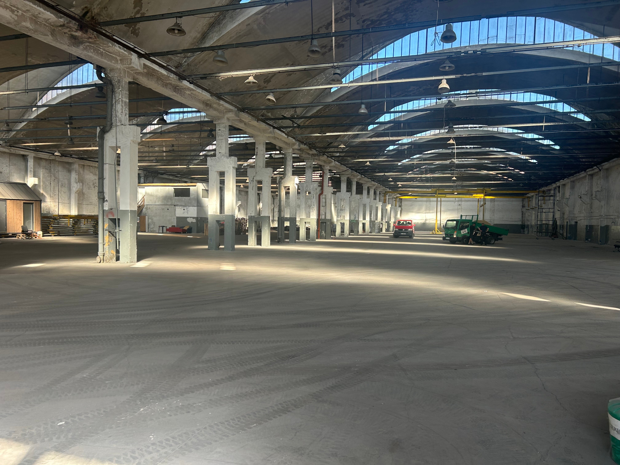 Przestronne wnętrze magazynu z betonową podłogą, rzędami kolumn i świetlikami w dachu. Widoczne ślady opon i pojazdy w oddali, podkreślające skalę i potencjał przestrzeni.