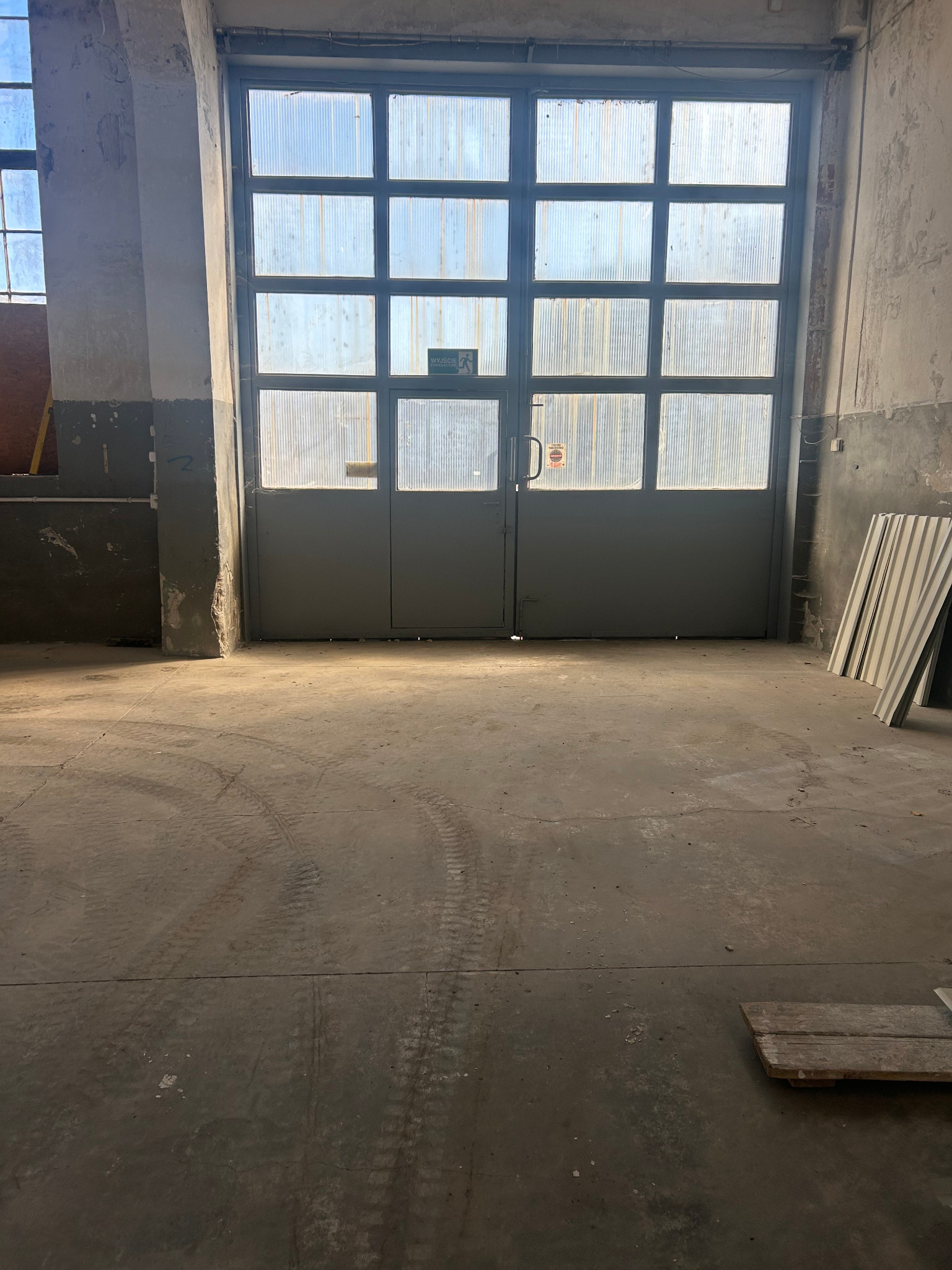 Wnętrze magazynu z betonową podłogą i dużymi, przeszklonymi drzwiami. Widoczne ślady opon na podłodze i fragmenty materiałów budowlanych przy ścianie.