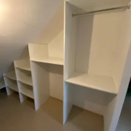 Biała szafa wnękowa na poddaszu ze skośnym sufitem, z półkami i drążkiem na ubrania, dopasowana do przestrzeni, na brązowych płytkach podłogowych.