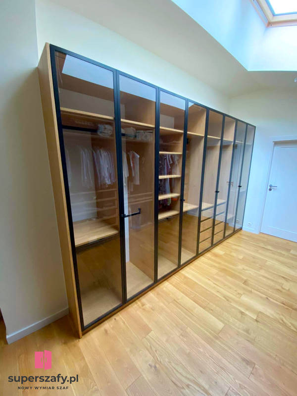 Nowoczesna szafa wnękowa z przeszklonymi drzwiami i czarnymi ramami, jasne drewno, podłoga z paneli, widok z dołu w kierunku okna dachowego.