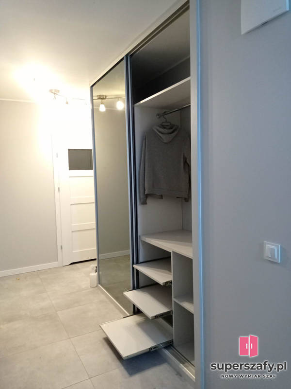 Szafa wnękowa z lustrzanymi drzwiami przesuwnymi i wysuwanymi półkami. W szafie wisi bluza z kapturem. Widoczna podłoga z szarych płytek i białe drzwi w tle.