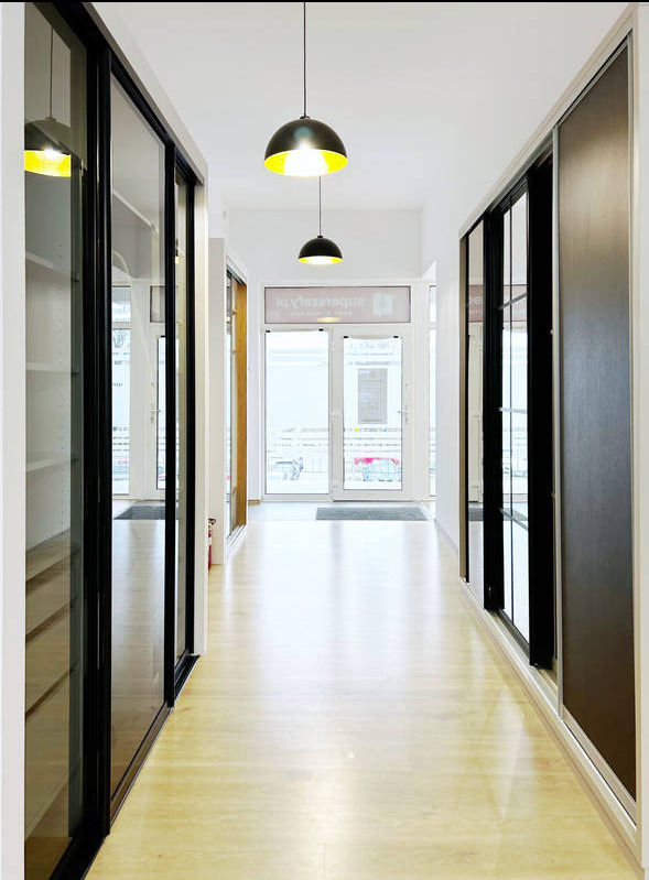 Długi korytarz z szafami wnękowymi po obu stronach, oświetlony wiszącymi lampami z żółtym światłem, z parkietem na podłodze i widokiem na zewnątrz przez przeszklone drzwi.