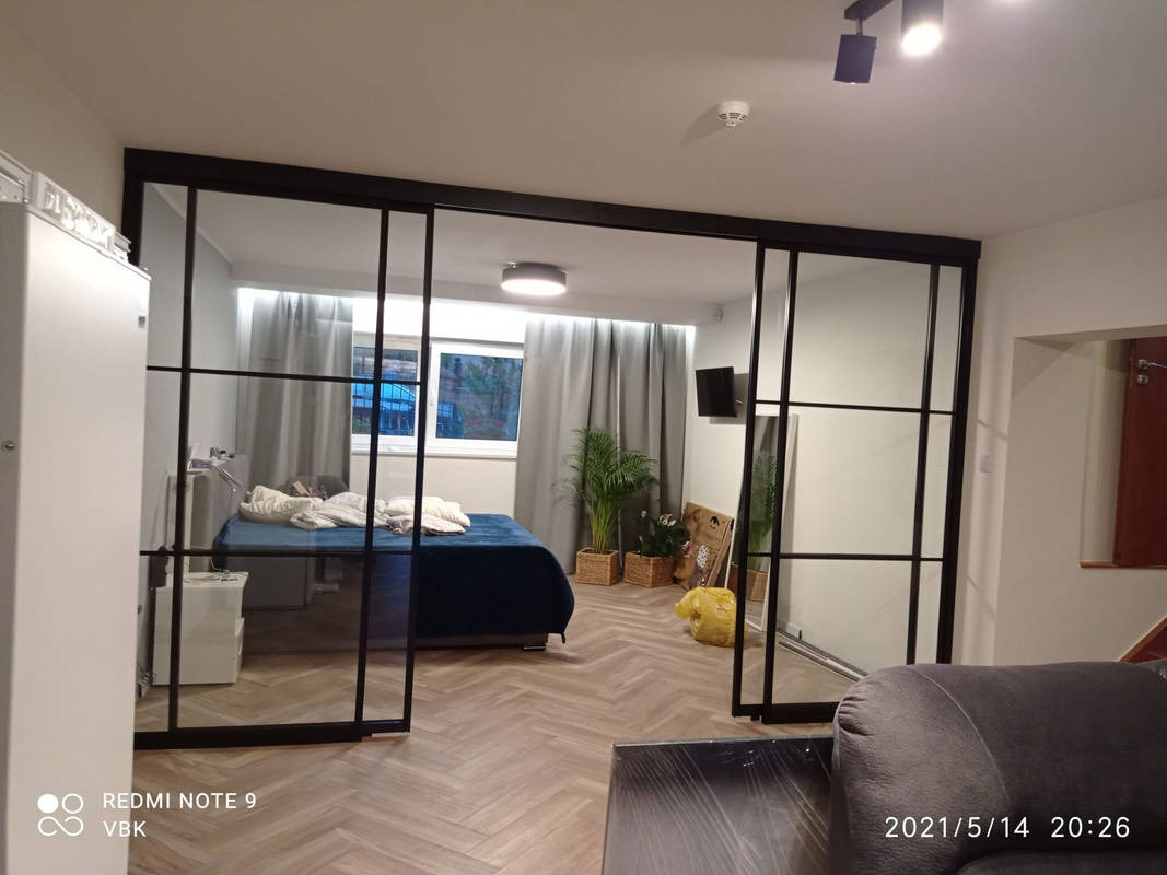 Nowoczesna szafa przesuwna z czarnymi szprosami oddzielająca sypialnię. Widoczny parkiet w jodełkę, łóżko i roślina w wiklinowym koszu. Minimalistyczny design.