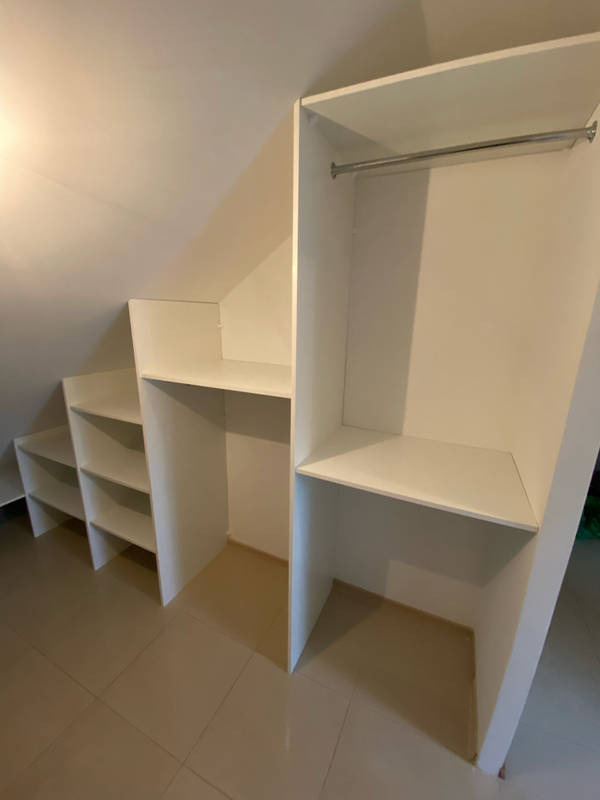 Biała szafa wnękowa na poddaszu ze skośnym sufitem, z półkami i drążkiem na ubrania, dopasowana do przestrzeni, na brązowych płytkach podłogowych.