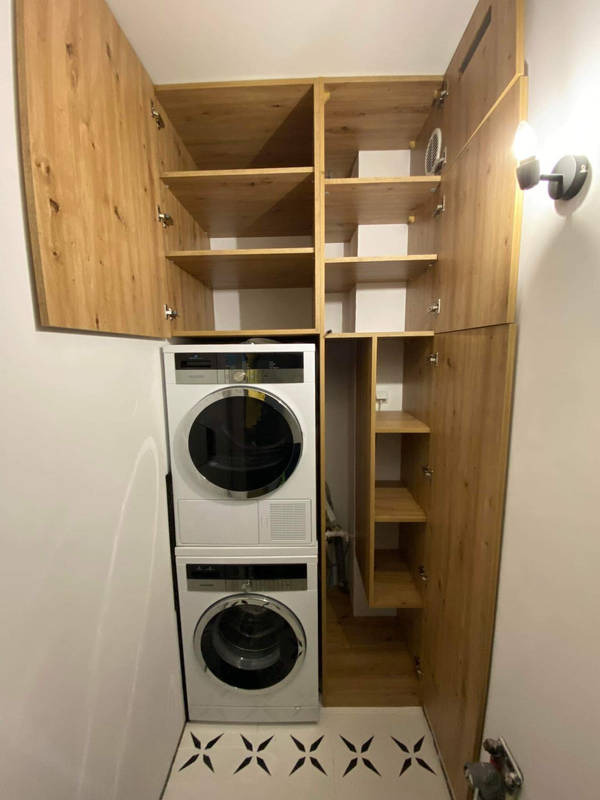 Drewniana szafa wnękowa z otwartymi drzwiami, w której znajduje się pralka i suszarka. Na podłodze wzorzyste płytki, oświetlenie boczne.