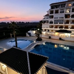 Edyta Bednarczyk - Apartamentowiec z basenami oświetlony wieczornym światłem. Widok z balkonu na kompleks wypoczynkowy i zachodzące słońce. Palmy i budynki w tle.