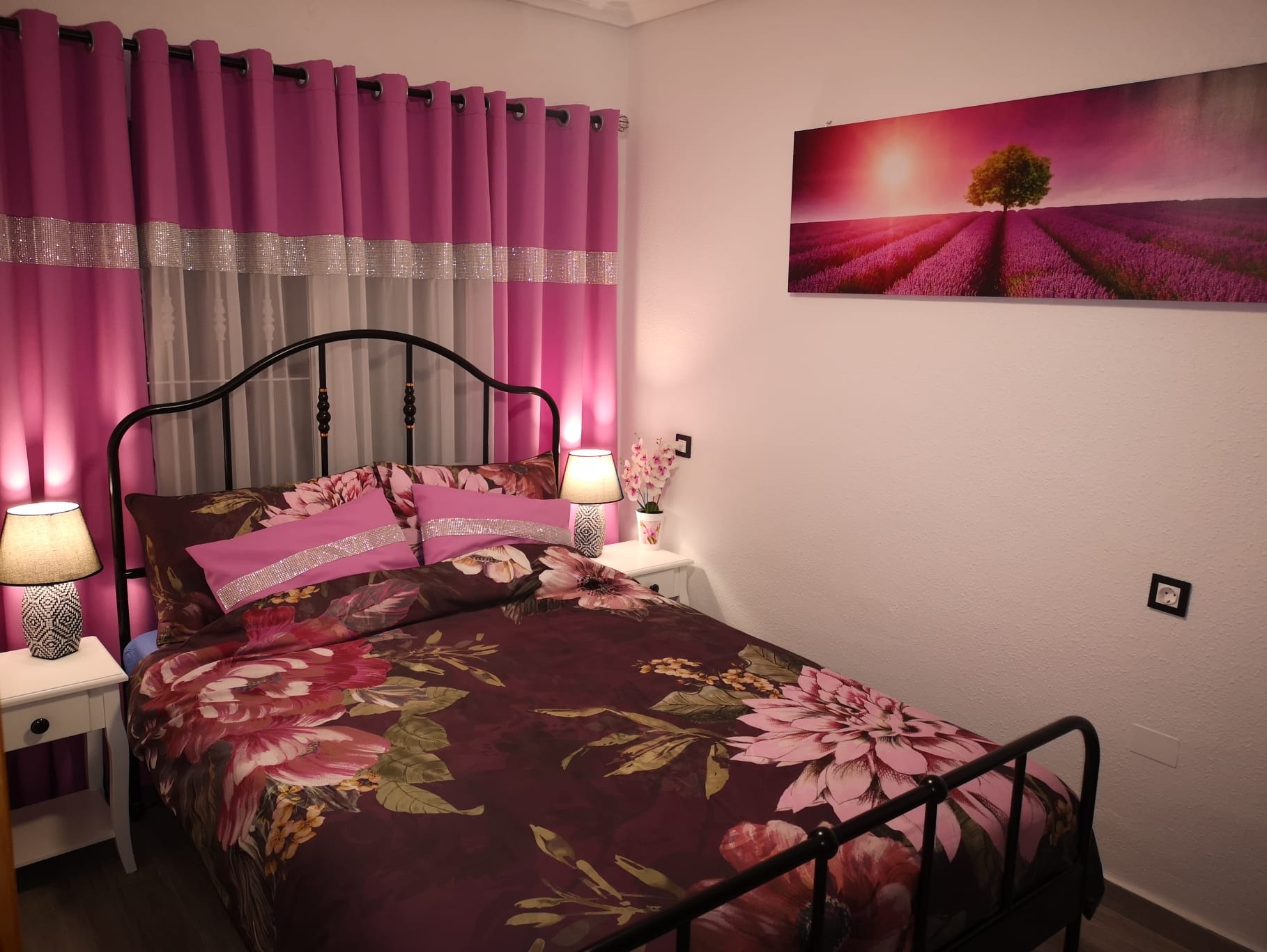 Przytulna sypialnia z łóżkiem w kwiaty, różowe zasłony z błyszczącymi paskami i obrazem lawendy. Dwie lampki nocne oświetlają wnętrze.
