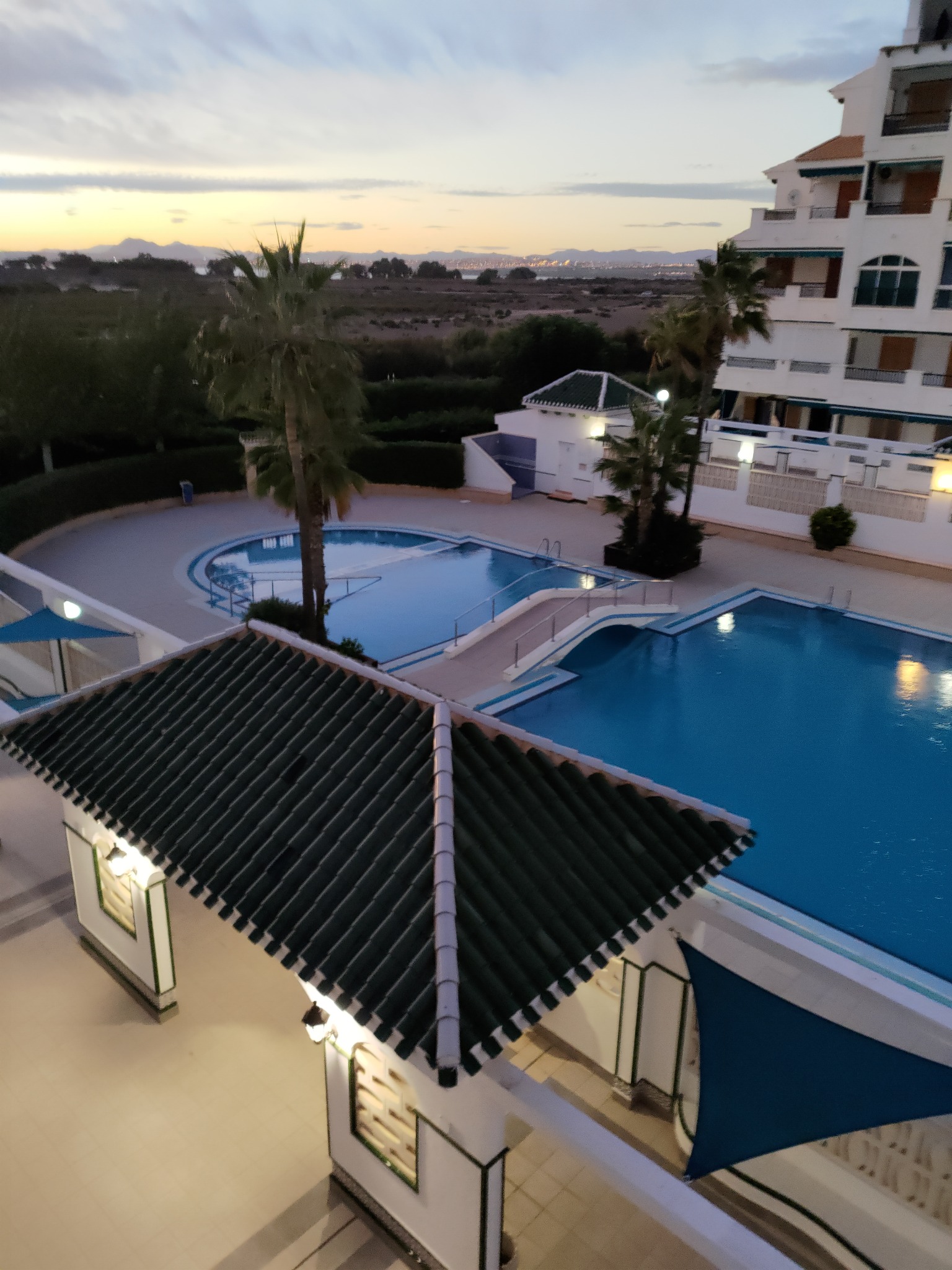 Kompleks basenowy z palmami o zmierzchu, widok z góry. Biały budynek w tle, zieleń w otoczeniu. Turkusowa woda w basenach, oświetlenie zewnętrzne.