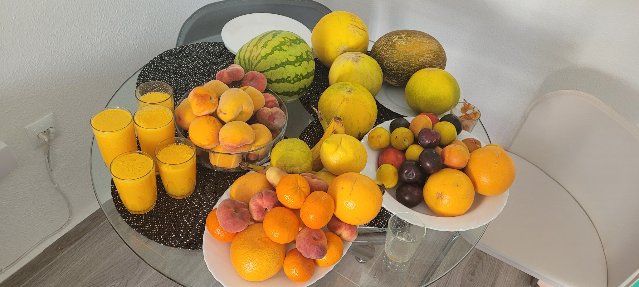 Szklany stół z owocami: arbuzy, melony, pomarańcze, brzoskwinie i śliwki. Obok szklanki ze świeżo wyciśniętym sokiem. Zdjęcie w jasnej tonacji, z gładkim tłem.