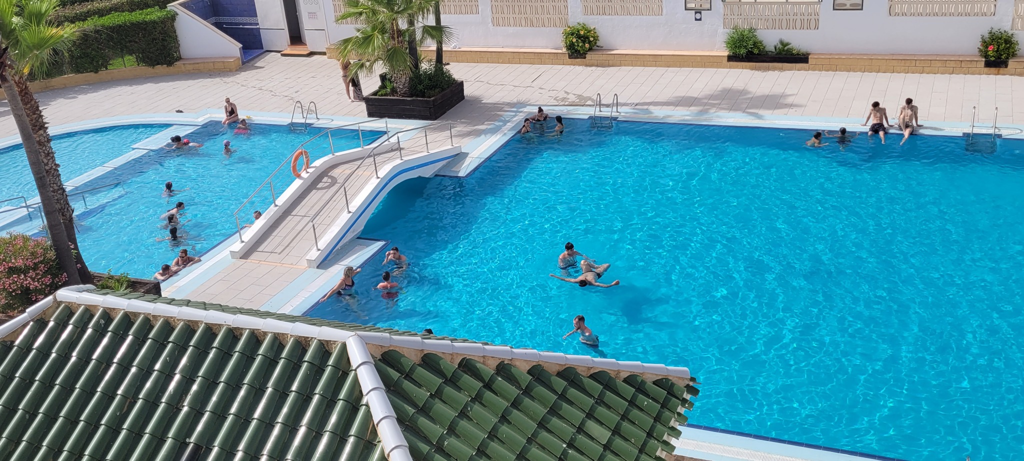 Widok z góry na basen z turkusową wodą, z mostkiem i palmami. W basenie pływają ludzie. W tle budynek z białymi ścianami i zielona roślinność.