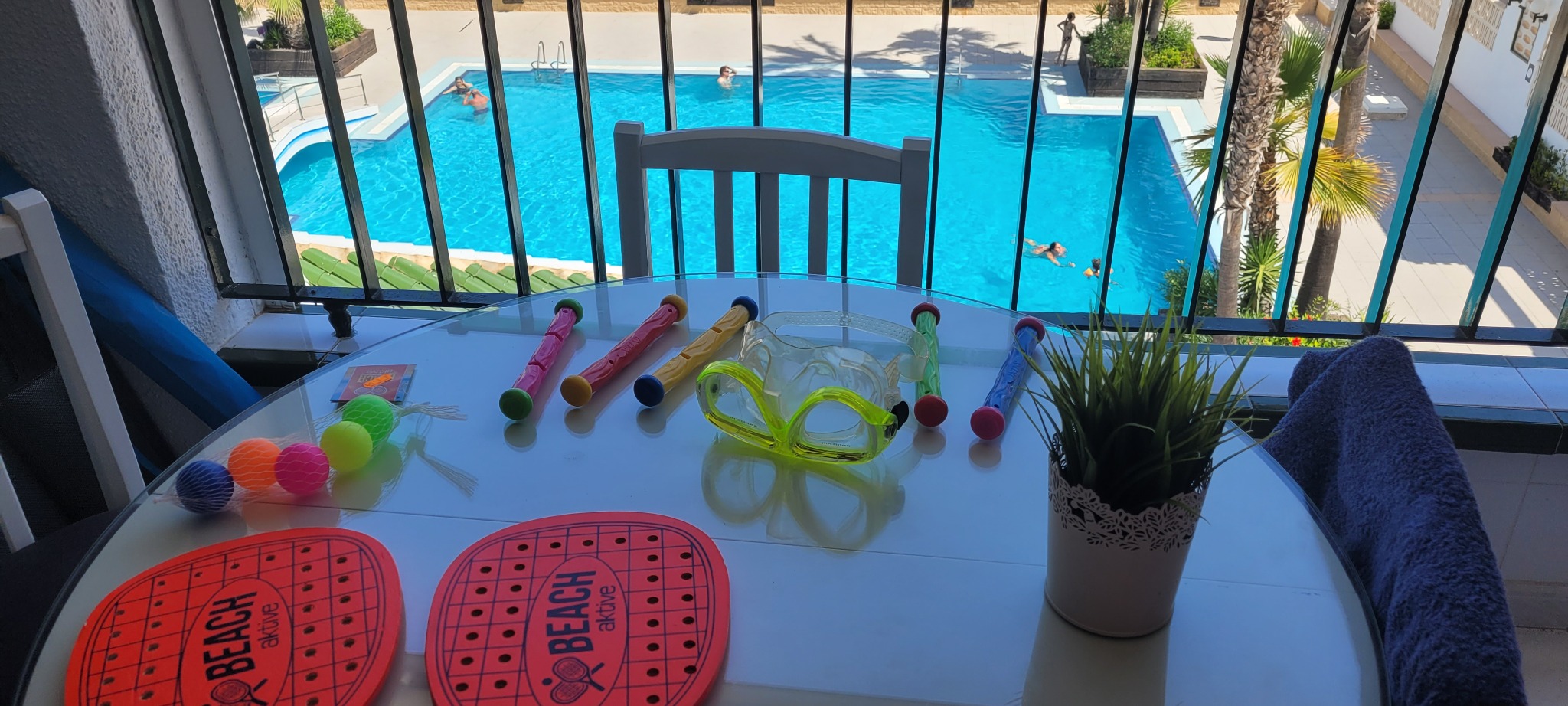 Balkon z widokiem na basen: rakietki plażowe, maska do nurkowania, zabawki wodne na stole. W tle osoby pływające w basenie.