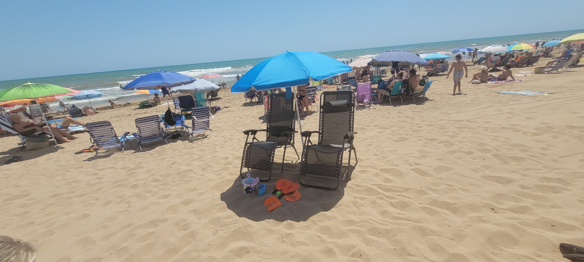 Pełna plaża z leżakami i parasolami, na pierwszym planie dwa czarne leżaki i zabawki dla dzieci. Błękitne niebo i morze w tle, słoneczny dzień.