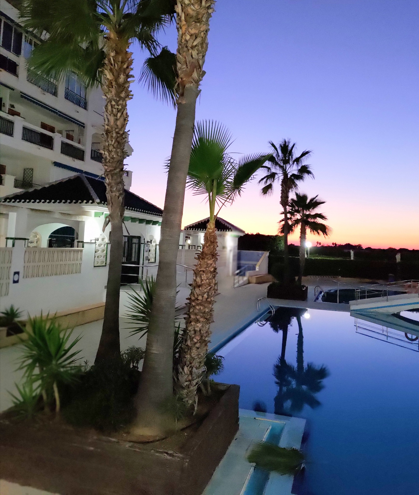 Błękitny basen z palmami o zmierzchu przy białym budynku z balkonami. Refleksy palm w wodzie. Krajobraz wakacyjny.