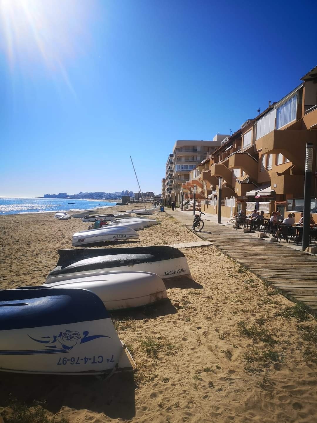 Słoneczny dzień na plaży: rzędy łodzi na piasku, budynki z balkonami w tle, ludzie siedzący w kawiarni na drewnianym chodniku wzdłuż plaży.