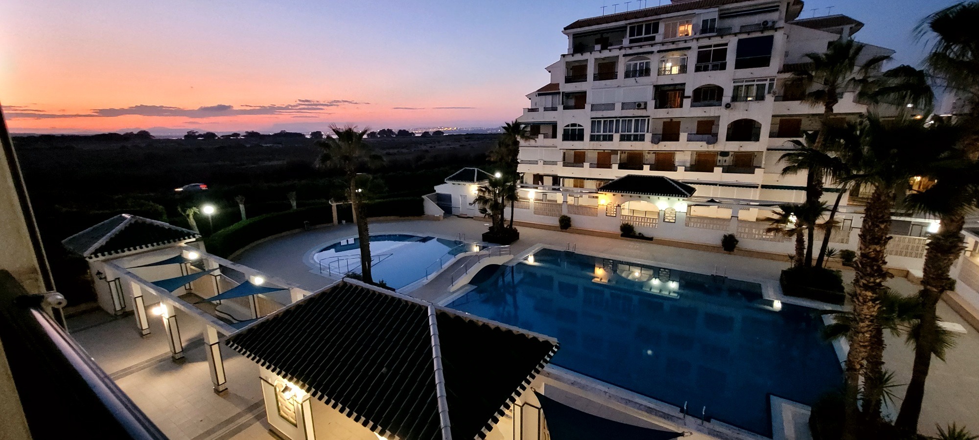 Apartamentowiec z basenami oświetlony wieczornym światłem. Widok z balkonu na kompleks wypoczynkowy i zachodzące słońce. Palmy i budynki w tle.