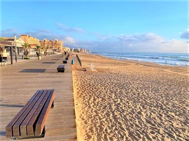 Słoneczny widok na plażę z drewnianym deptakiem i ławkami. W oddali budynki i spokojne morze pod błękitnym niebem.