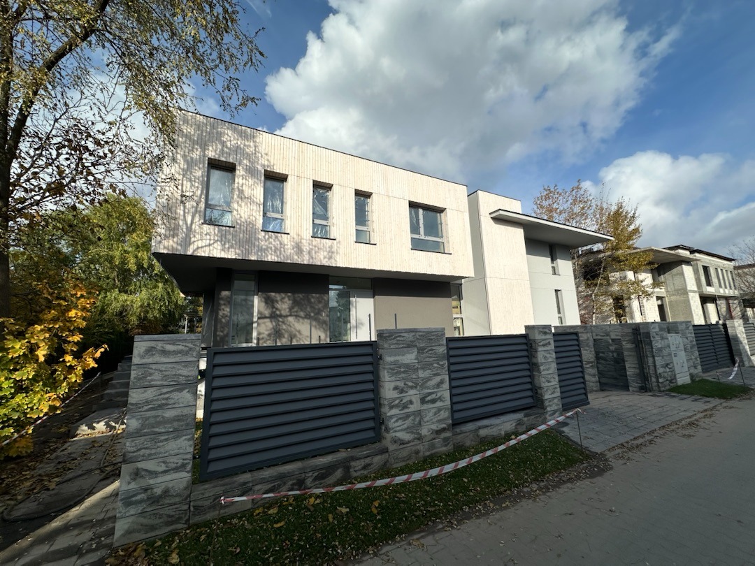 Nowoczesny dom z drewnianą elewacją i szarym ogrodzeniem panelowym, widok z poziomu ulicy w słoneczny dzień. Widoczne drzewa i inne budynki.