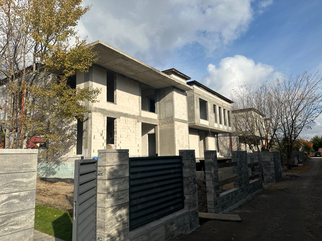 Surowy stan domu z betonu komórkowego, widok z zewnątrz, ogrodzenie panelowe z betonowych elementów, jesienne drzewa, błękitne niebo z chmurami w tle.