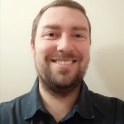 Uśmiechnięty portret mężczyzny z brązowymi włosami i brodą, ubranego w ciemną koszulę, na jasnym tle. Widoczne detale twarzy i górnej części ciała.