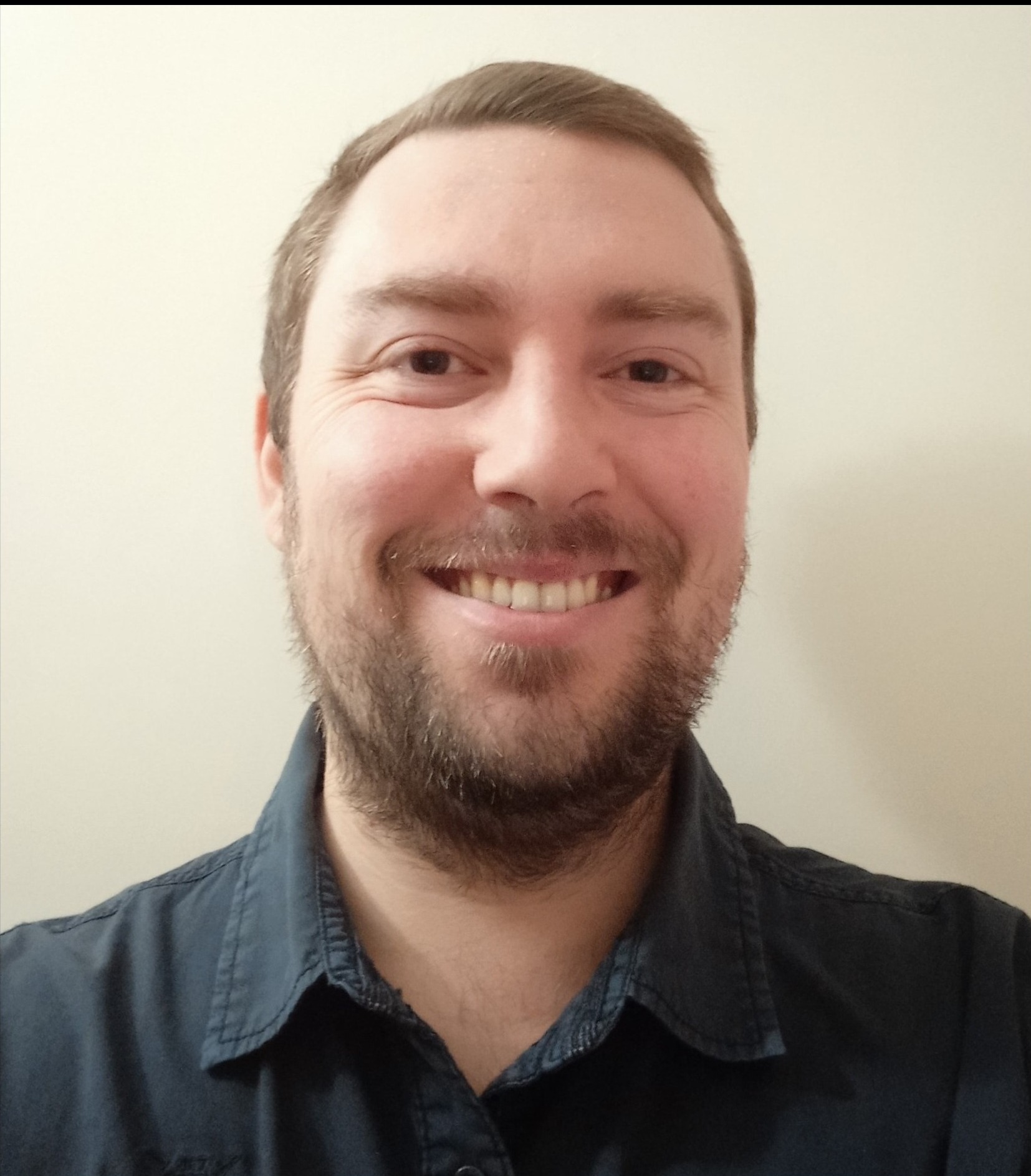 Uśmiechnięty portret mężczyzny z brązowymi włosami i brodą, ubranego w ciemną koszulę, na jasnym tle. Widoczne detale twarzy i górnej części ciała.