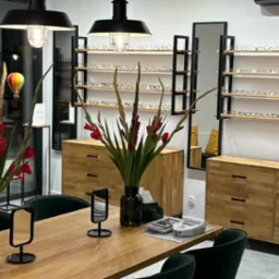 Wnętrze salonu optycznego: drewniany stół z kwiatami, ekspozycja okularów na półkach, komody z drewna. Dekoracje balonowe dodają uroku.