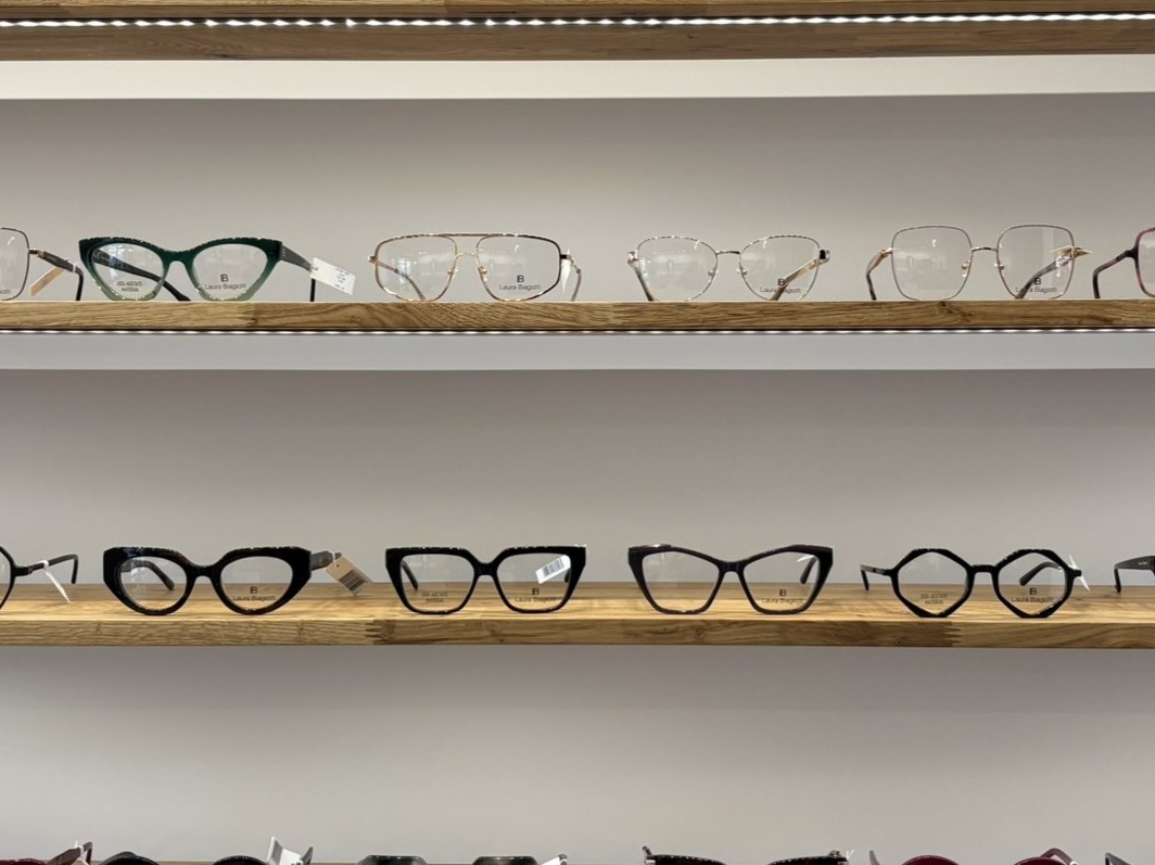 Różnorodne okulary korekcyjne marki Laura Biagiotti, ułożone na drewnianych półkach w salonie optycznym. Widoczne modele w kolorach czarnym, złotym i zielonym.