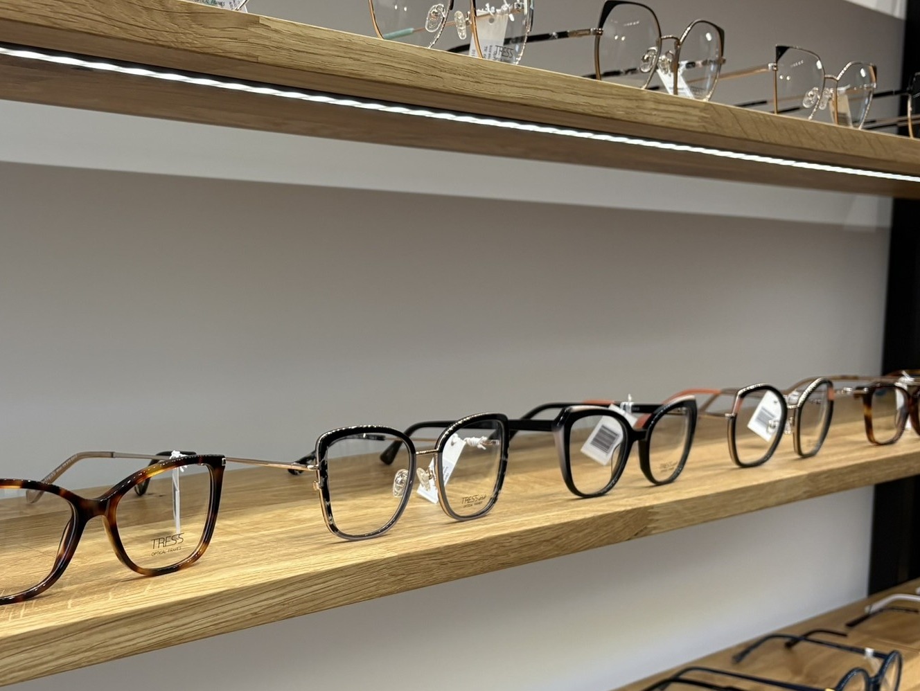 Różnorodne okulary korekcyjne marki Tress, starannie ułożone na drewnianych półkach w salonie optycznym, oświetlone subtelnym światłem LED. Prezentacja asortymentu.