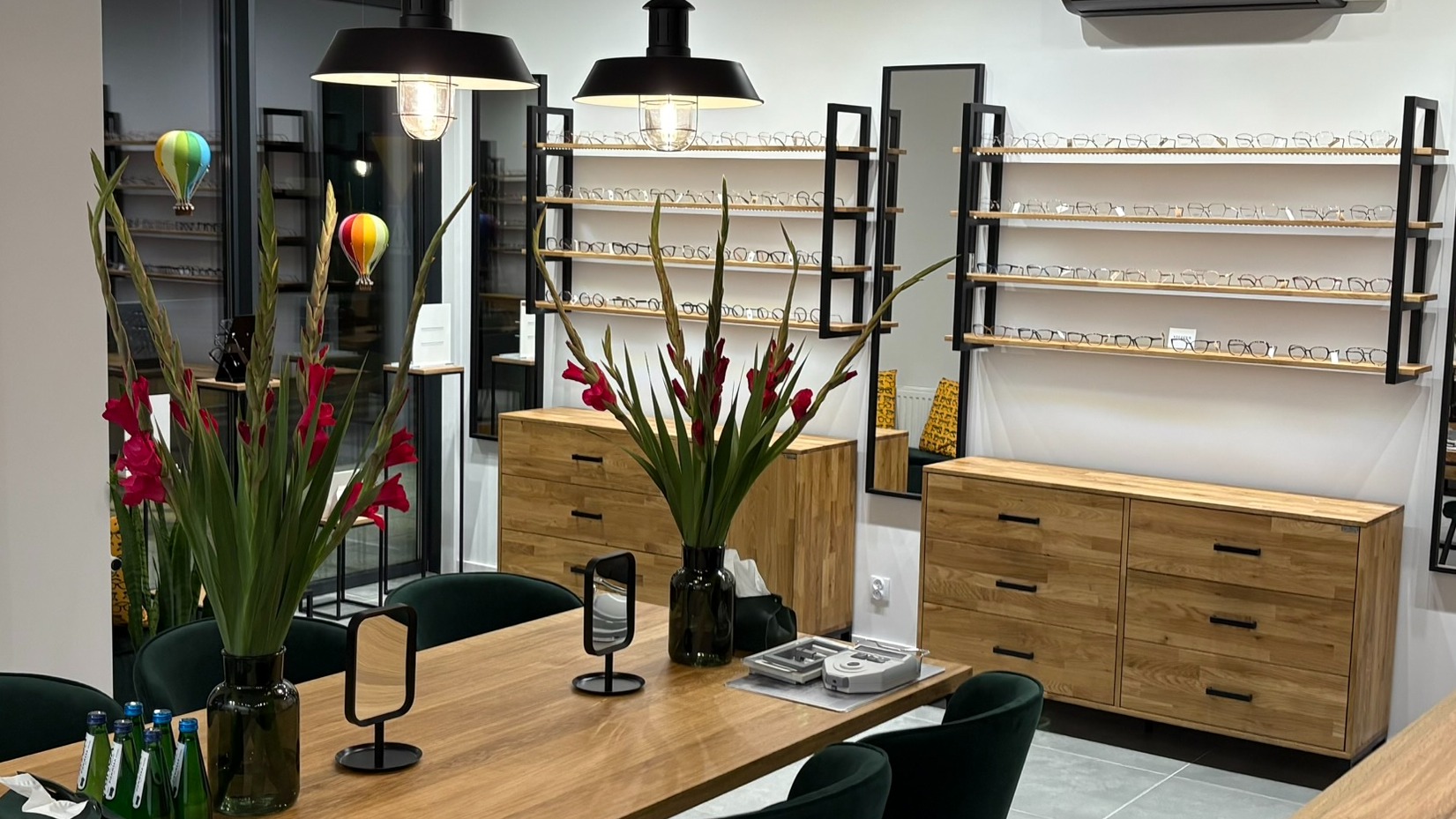 Wnętrze salonu optycznego: drewniany stół z kwiatami, ekspozycja okularów na półkach, komody z drewna. Dekoracje balonowe dodają uroku.