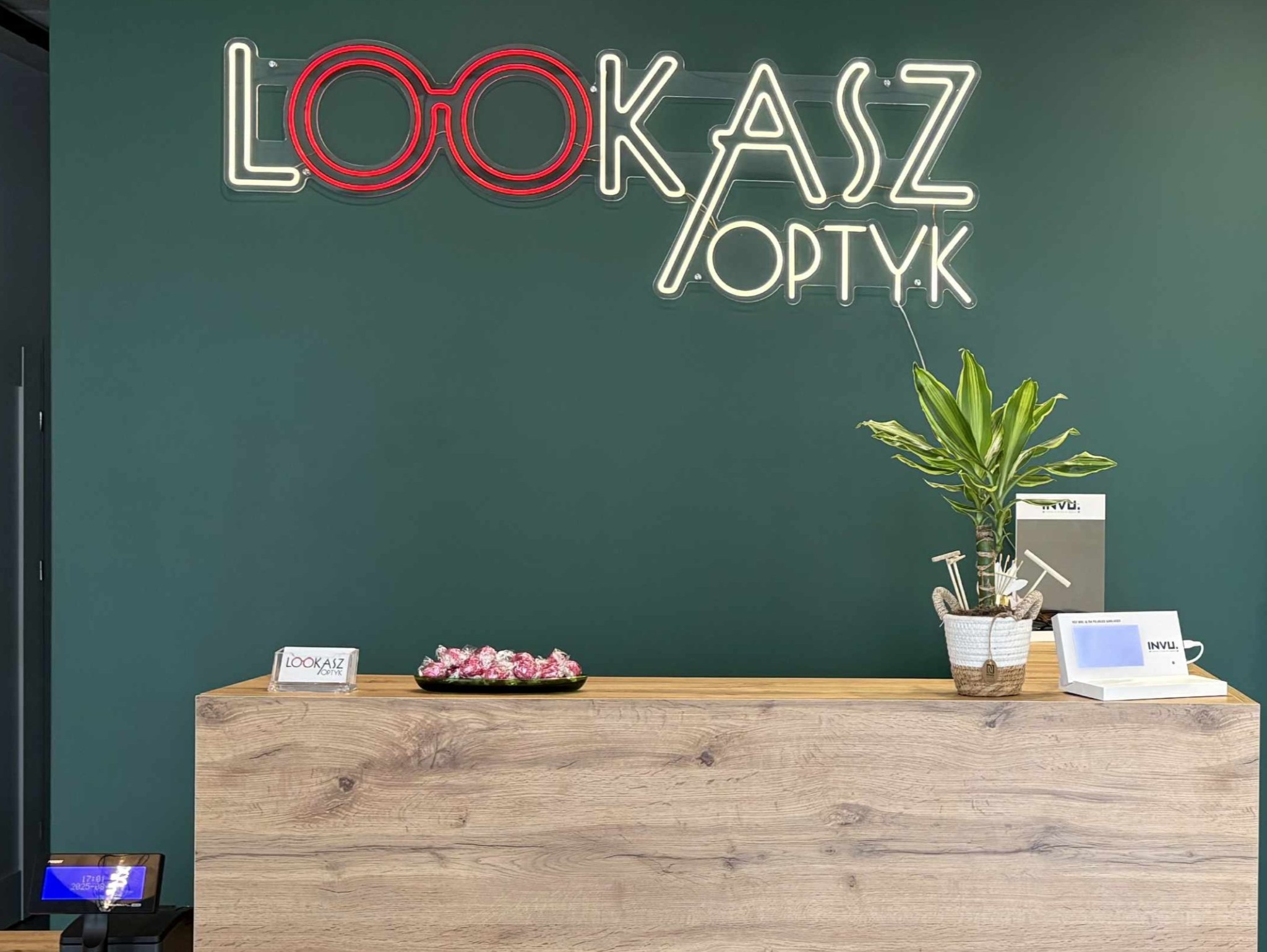 Recepcja w salonie optycznym z neonowym logo 'Lookasz Optyk', zieloną ścianą, drewnianą ladą i elementami dekoracyjnymi.