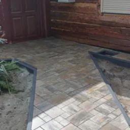 Ułożona kostka brukowa na podjeździe drewnianego domu z szarą ramą obrzeża. Widoczna brązowa stolarka okienna i drzwiowa oraz skrzynka elektryczna na elewacji.