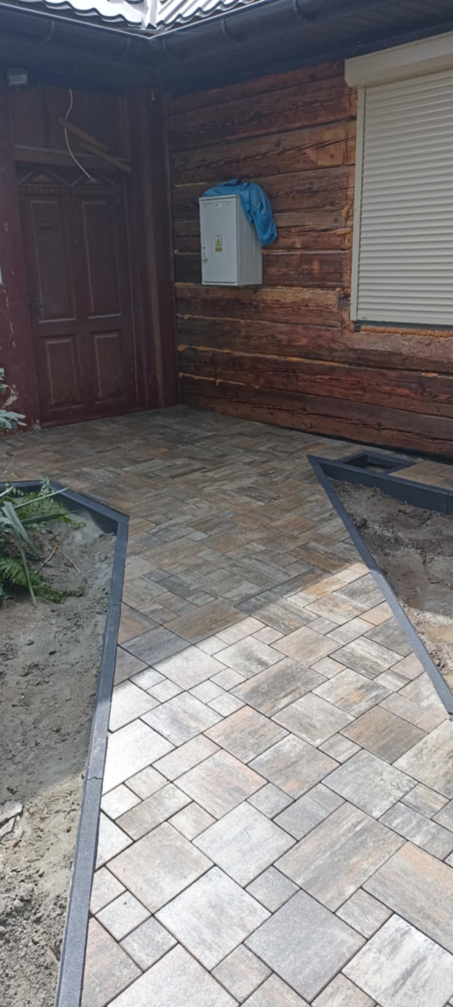 Ułożona kostka brukowa na podjeździe drewnianego domu z szarą ramą obrzeża. Widoczna brązowa stolarka okienna i drzwiowa oraz skrzynka elektryczna na elewacji.