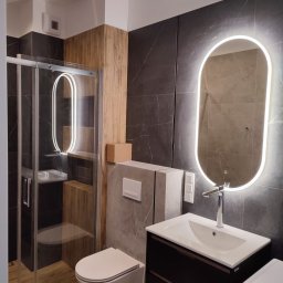 Masterscraft Renovations sp z o.o - Nowoczesna łazienka po remoncie: kabina prysznicowa ze szklanymi drzwiami, podwieszana toaleta, umywalka z szafką i podświetlane lustro owalne na tle grafitowych płytek.