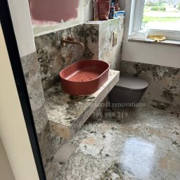 Masterscraft Renovations sp z o.o - Wykończona łazienka z umywalką w kolorze terakoty i armaturą w odcieniu mosiądzu. Na ścianach i podłodze płytki imitujące kamień. Widoczne materiały budowlane.