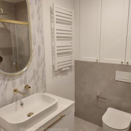 Masterscraft Renovations sp z o.o - Nowoczesna łazienka z owalnym lustrem w złotej ramie, białą umywalką i grzejnikiem drabinkowym. Uwagę zwracają płytki w kształcie strzałek i złota armatura.