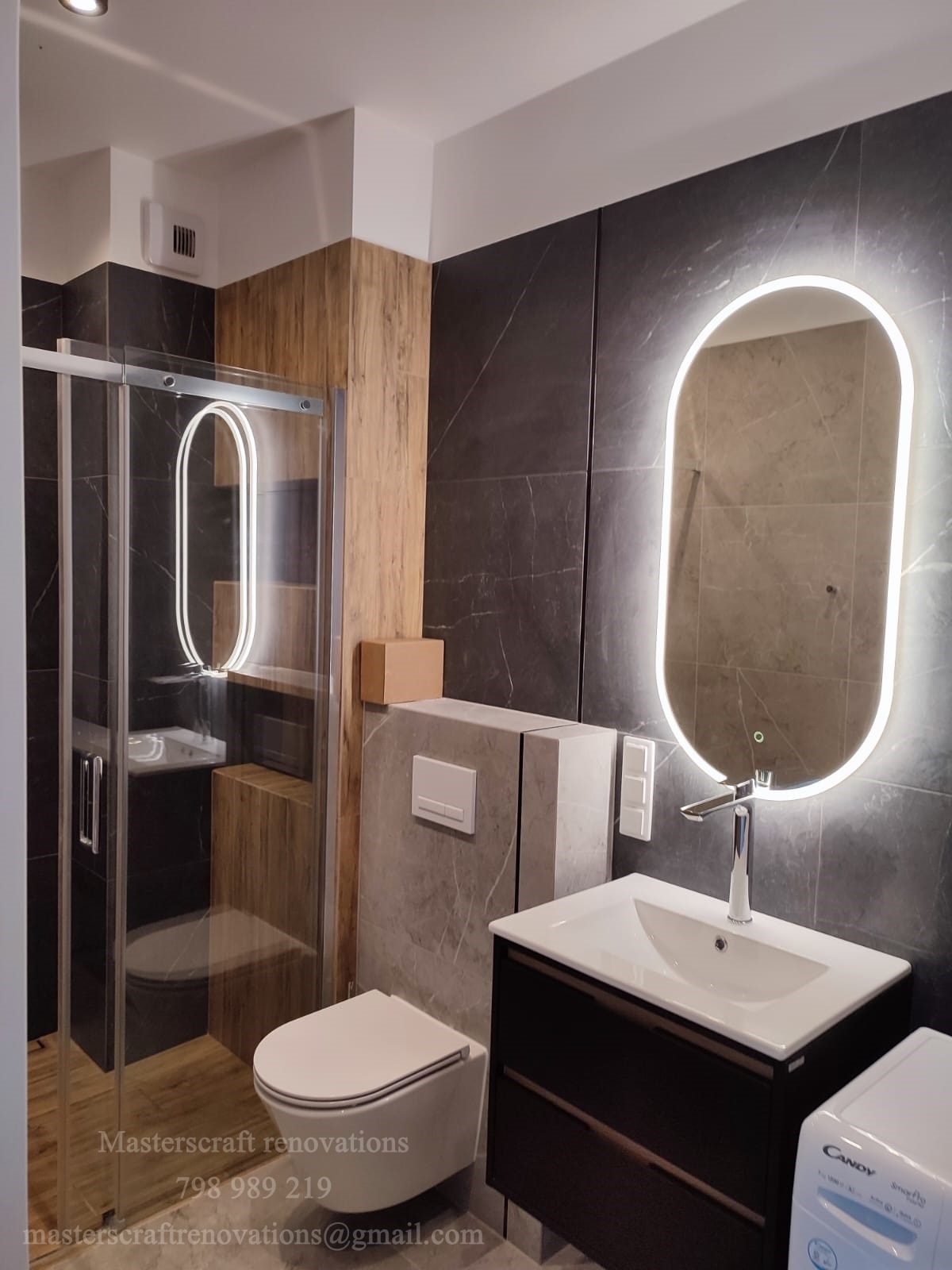 Nowoczesna łazienka po remoncie: kabina prysznicowa ze szklanymi drzwiami, podwieszana toaleta, umywalka z szafką i podświetlane lustro owalne na tle grafitowych płytek.