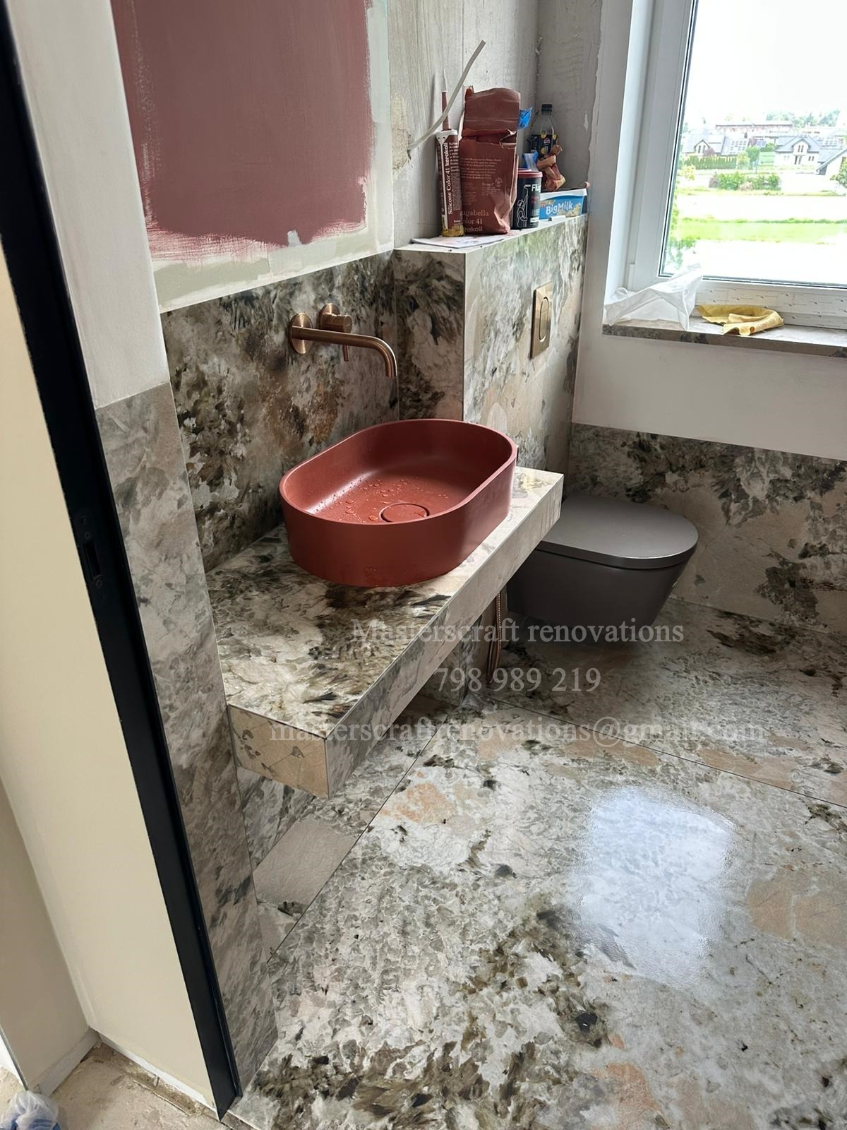 Wykończona łazienka z umywalką w kolorze terakoty i armaturą w odcieniu mosiądzu. Na ścianach i podłodze płytki imitujące kamień. Widoczne materiały budowlane.