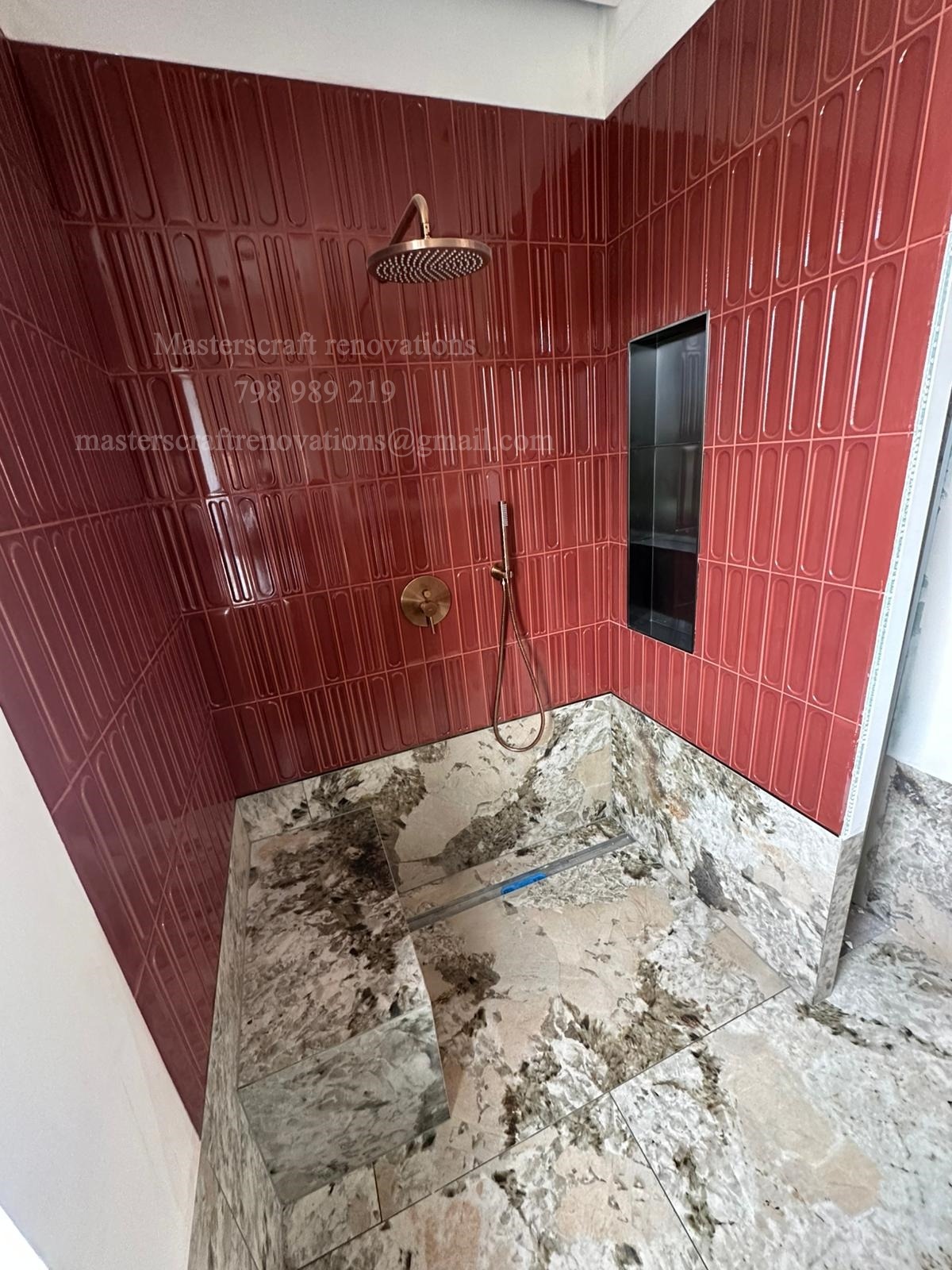 Nowoczesna kabina prysznicowa z bordowymi, podłużnymi płytkami i kamienną posadzką. Widoczna deszczownica i wnęka w ścianie.