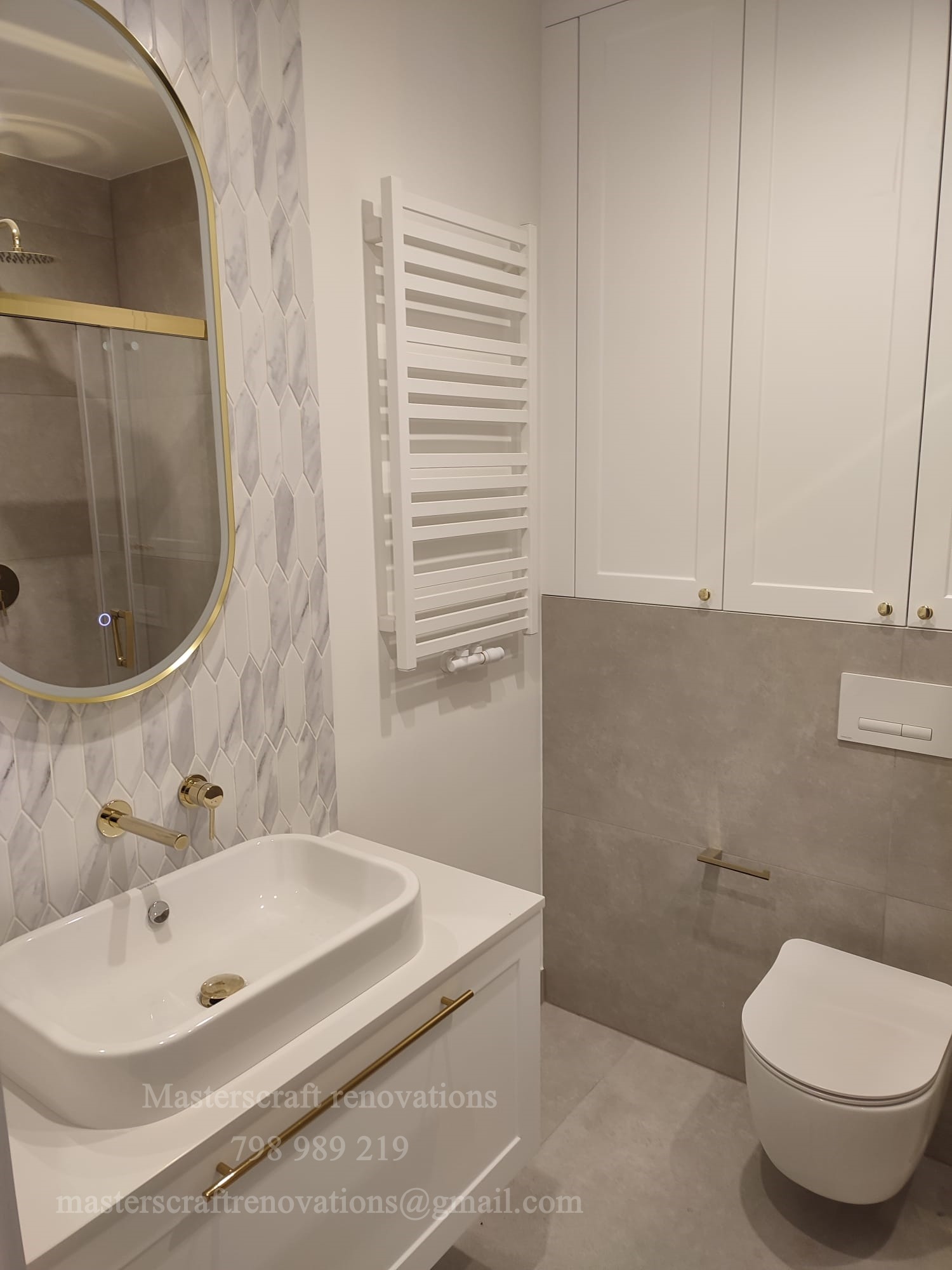 Nowoczesna łazienka z owalnym lustrem w złotej ramie, białą umywalką i grzejnikiem drabinkowym. Uwagę zwracają płytki w kształcie strzałek i złota armatura.