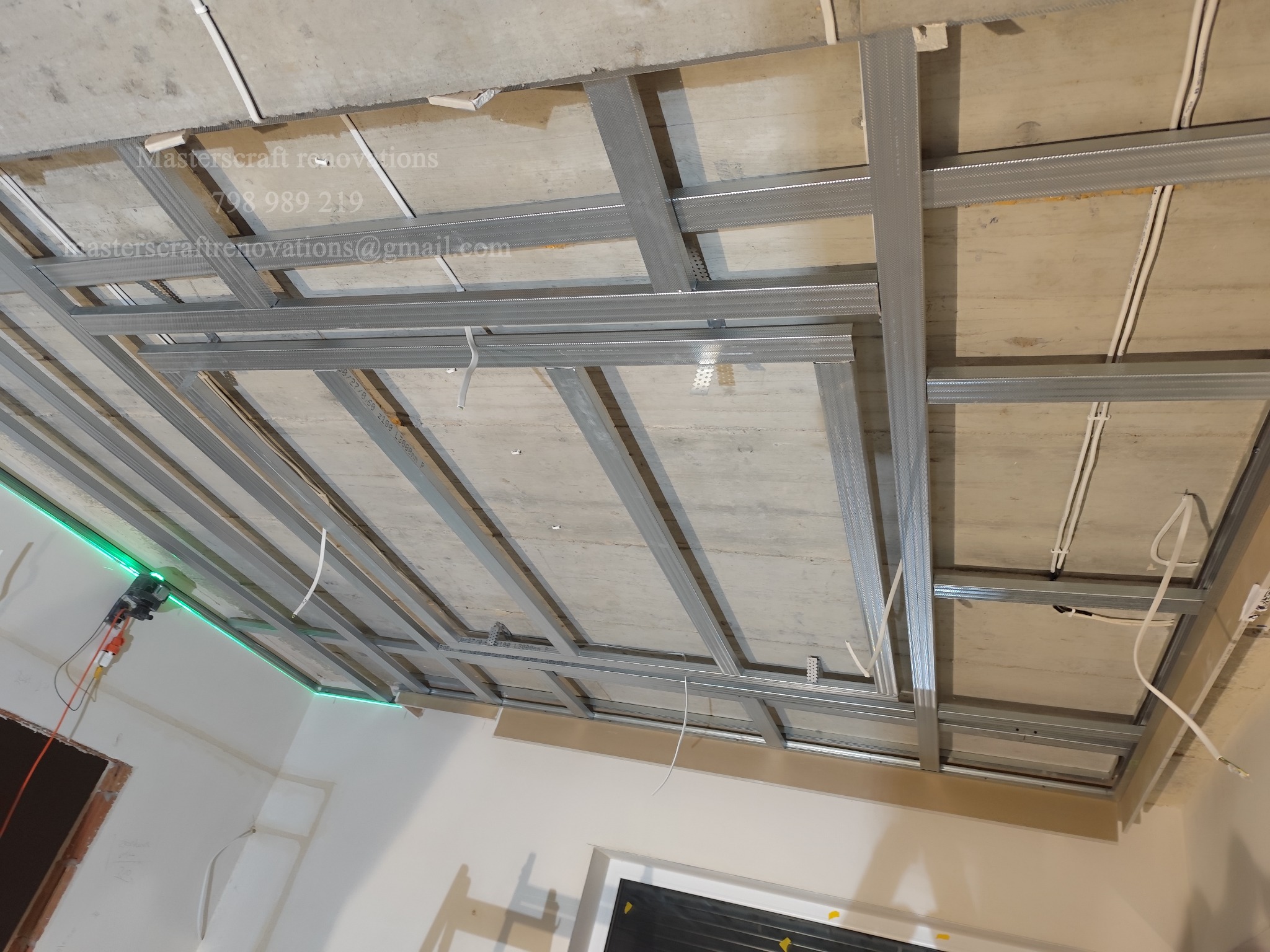 Metalowa konstrukcja sufitu podwieszanego, widok od dołu. Widoczne profile, przewody instalacji elektrycznej i fragment ściany z zielonym oświetleniem.