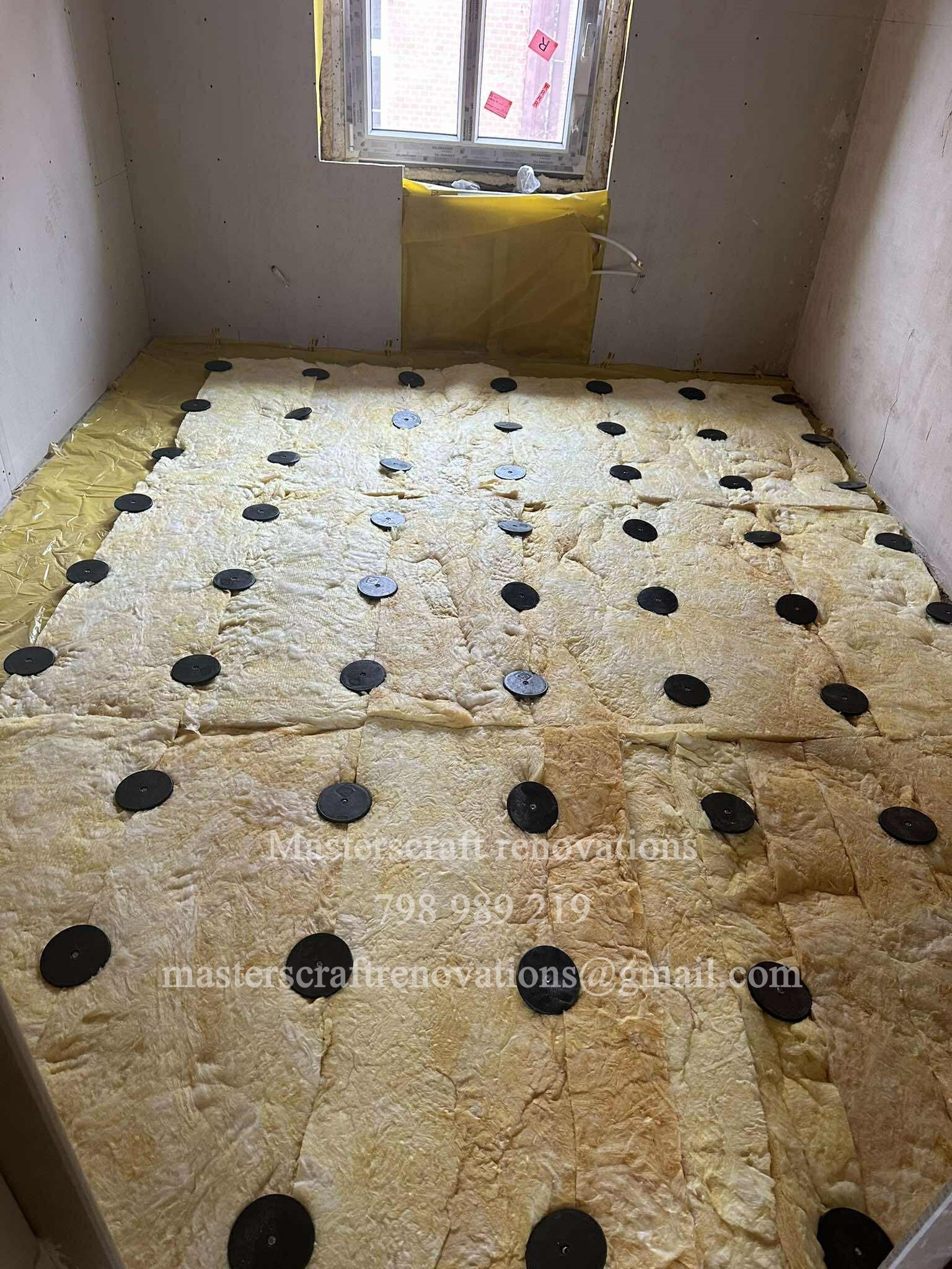 Ocieplenie podłogi wełną mineralną z czarnymi uchwytami mocującymi, przygotowane do wylewki. Widoczne fragmenty ścian i okno w tle.