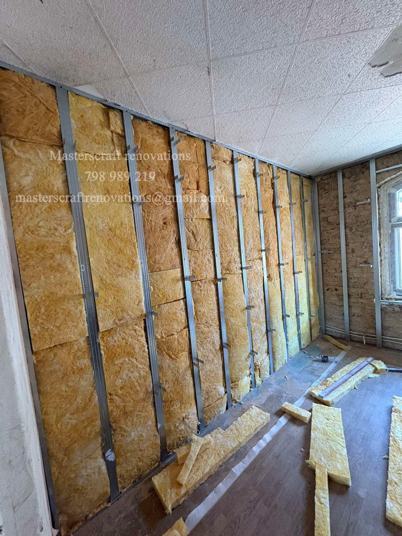 Ściana w trakcie izolacji termicznej: wełna mineralna między metalowymi profilami, widoczne fragmenty instalacji i okno. Etap prac wykończeniowych.